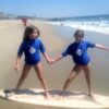 2 girls holding hands on a surfboard on the beach in Los Angeles